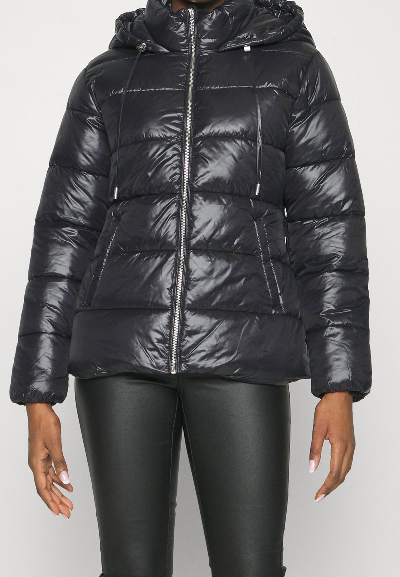 Femme portant une doudoune matelassée noire brillante avec capuche et fermeture éclair frontale, associée à un pantalon en cuir noir ajusté.