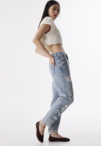 Häkelgestricktes cremefarbenes Crop-Top, kombiniert mit hellblauen, zerrissenen Jeans mit schwarzem grafischem Text. Das Model trägt braune Slipper mit Kettendetail.