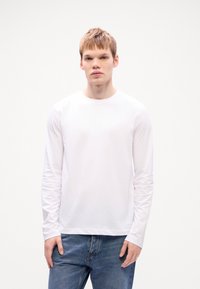 Balta ilga rankovė t-shirt, pagaminta iš minkštos medvilnės. Paprastas apvalios apykaklės dizainas su tiesia apačia. Suderinta su mėlynais džinsais.