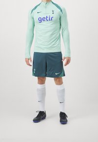 Minttiharmaa pitkähihaisen jalkapallopaidan violetti logo, yhdistettynä tummansinisiin shortseihin, valkoisiin sukkiin ja mustiin nappulakenkiin, joissa on sinisiä yksityiskohtia.