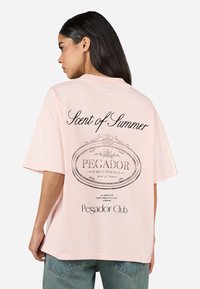 Lys rosa bomull T-skjorte med avslappet passform. Har svart trykt tekst på baksiden: "Duft av Sommer" og "Pegador Club" i et sirkulært design.