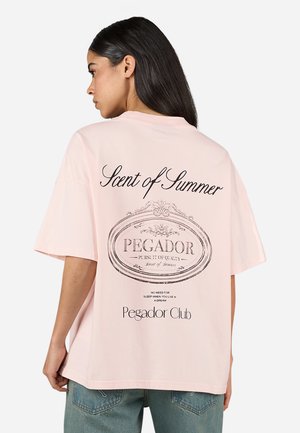 Lichtroze katoenen T-shirt met een losse pasvorm. Heeft zwarte bedrukte tekst op de achterkant: "Geurovereenstemming van de Zomer" en "Pegador Club" in een circulair ontwerp.