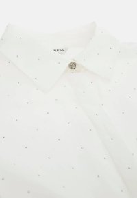 Camicia bianca con colletto, caratterizzata da strass argentati sparsi sul tessuto e un bottone logo rotondo argento al collo.