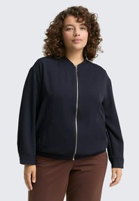 Veste de bombardier marine avec fermeture éclair à l'avant, col côtelé, poches latérales et coupe décontractée ; présente une texture lisse et un ourlet légèrement élastique.