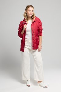 Jeune femme portant une veste rouge sur un haut blanc, un pantalon large blanc et des chaussures ouvertes à talons blancs, sur un fond uni.