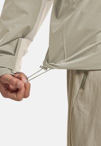 Mano che regola il cordoncino dell'orlo di una giacca beige, mostrando il polsino della manica con strap in Velcro e la tasca di pantaloni beige con zip.