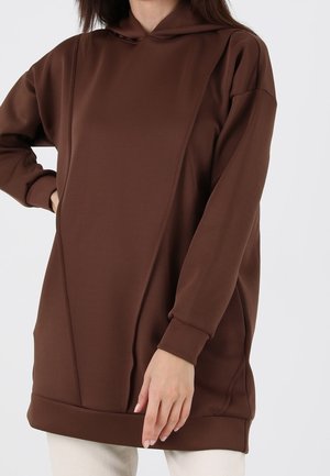 Femme portant un sweat à capuche marron uni, oversize, avec des poches, debout devant un fond clair.