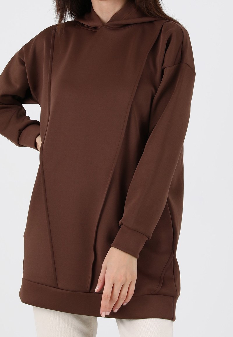 Femme portant un sweat à capuche marron uni, oversize, avec des poches, debout devant un fond clair.