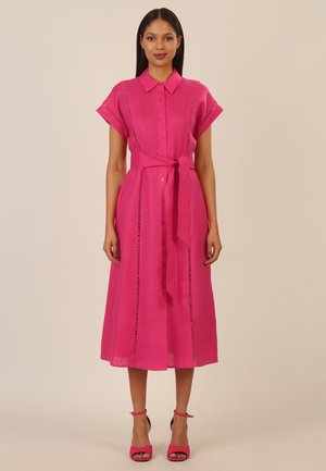 Frau steht und trägt ein hellrosa Midikleid mit kurzen Ärmeln, Knopfleiste vorne, Taillengürtel und pinken Sandalen mit hohen Absätzen.