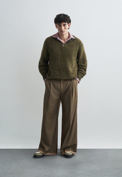 Olivgrüner strukturierter Pullover mit Reißverschlusskragen, über einem rosa Hemd mit Kragen getragen. Locker sitzende braune Hose und zweifarbige Schuhe.