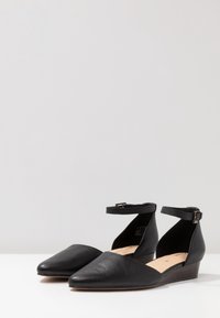 Chaussures noires en cuir avec une bride à la cheville, à bout pointu et talon compensé bas, affichant une texture lisse et un design minimaliste.