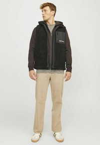 Chaleco de forro polar negro con capucha, cremallera frontal y bolsillos laterales, superpuesto sobre una camiseta de manga larga oscura y una camiseta gris, combinado con pantalones beiges.