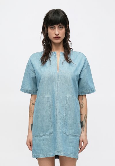 Kvinna med mörkt hår och tatueringar, klädd i en lös, kortärmad ljusblå denimklänning med framfickor, stående mot en enfärgad vit bakgrund.