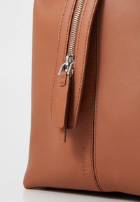 Sac en cuir marron avec une texture lisse, doté d'une fermeture éclair argentée et d'une forme rectangulaire. Comprend un tirette en cuir et un branding subtil.