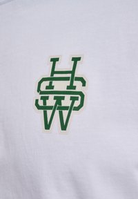 Baltas medvilnės audinys su žaliu ir kreminiu siuvinėtu logotipu, kuriame susipina raidės "H", "S", "W". Dizainas yra centrinis ir ryškus.