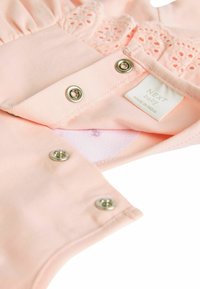 Helles rosa Babybekleidungsstück mit strukturiertem Spitze-Detail, silbernen Druckknöpfen und einem Etikett mit der Aufschrift "NEXT baby", hergestellt in Indien.