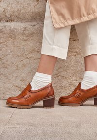 Brown leather block-heeled shoes with a rounded toe, decorative stitching, and a chain accent. Worn with white ribbed socks.