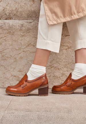 Brown leather block-heeled shoes with a rounded toe, decorative stitching, and a chain accent. Worn with white ribbed socks.