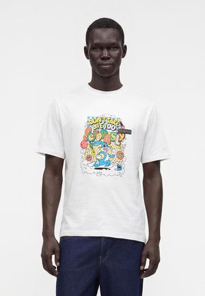 Junger Mann trägt ein weißes T-Shirt mit bunten Cartoon-Grafiken und dem Schriftzug "Nimm das Leben nicht zu ernst", kombiniert mit dunkelblauen Jeans.