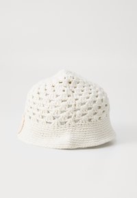 Háčkovaná čiapka v off-white farbe, ktorá má štruktúrovaný otvorený vzor a jemne rozšírený okraj. Doplnená malou nášivkou s logom.