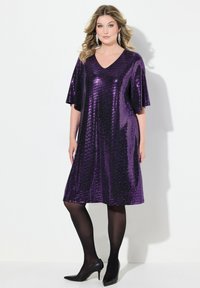 Robe violette à paillettes avec décolleté en V et manches courtes, présentant une coupe ample qui descend jusqu'à mi-mollet, assortie à des collants noirs transparents et des talons.