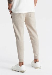 Beige sweatpants met een tapered fit, gemaakt van zachte stof; voorzien van elastische boorden, een achterzak en logo-detail. G gedragen met witte sneakers.