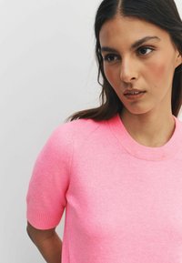 Pulover z kratkimi rokavi, okrogel izrez, svetlo roza barve. Gladek material z rebrastimi manšetami. Povečana slika poudarja izrez in detajle na ramenu.