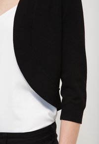 Černý zkrácený kardigan s tříčtvrtečními rukávy, vyrobený z měkké látky. Má zaoblený přední design a hladkou texturu, nošený přes bílý top.