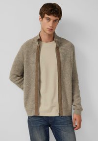Beige gebreide cardigan met een textuurpatroon, rits aan de voorkant en bruine afwerking. Gecombineerd met een lichtgekleurde t-shirt en blauwe jeans.