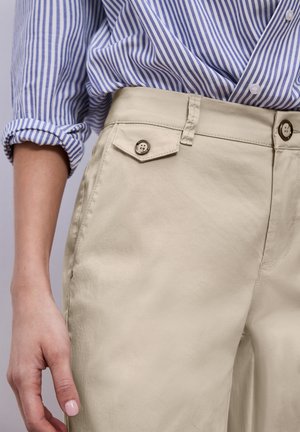 Donna che indossa pantaloni beige con pattina della tasca con bottoni e camicia a righe blu e bianche con maniche arrotolate.