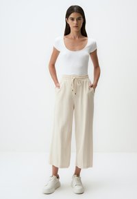 Witte geribbelde fitted top, beige wijde broek met een elastische tailleband en zakken, gecombineerd met witte sneakers op een effen achtergrond.