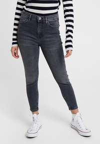 Osoba ubrana w czarne, przetarte skinny jeans, długi czarny i biały pasiasty top oraz białe trampki typu high-top stoi.