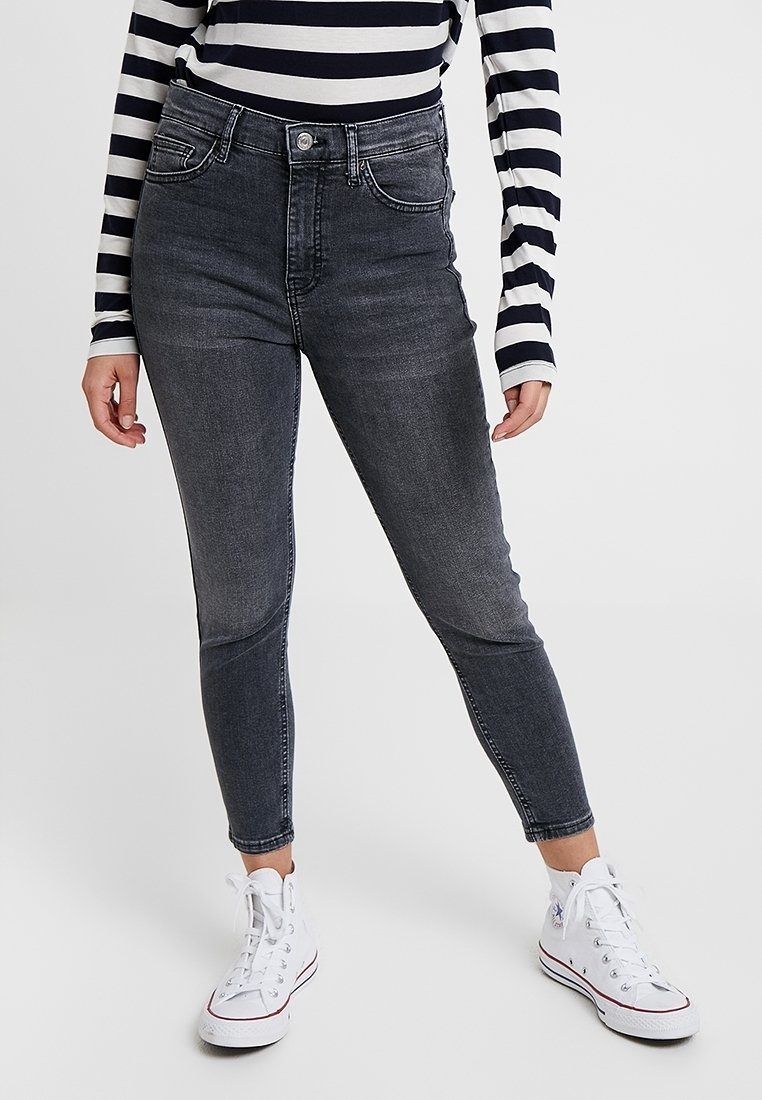 Osoba ubrana w czarne, przetarte skinny jeans, długi czarny i biały pasiasty top oraz białe trampki typu high-top stoi.