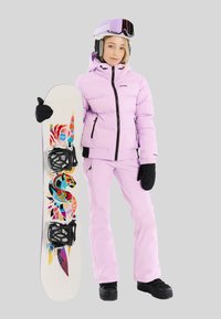 Jonge vrouw in een roze skikleding en helm, die een witte snowboard vasthoudt met kleurrijke abstracte patronen op een grijze achtergrond.