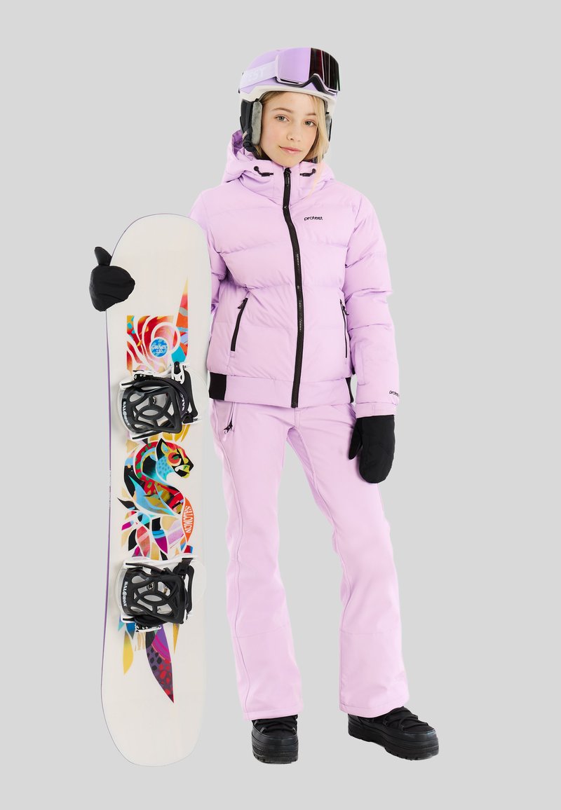 Jonge vrouw in een roze skikleding en helm, die een witte snowboard vasthoudt met kleurrijke abstracte patronen op een grijze achtergrond.