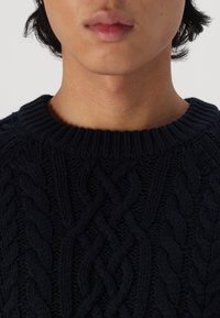 Marineblauer Strickpullover mit Kabelmuster, rundem Ausschnitt und geripptem Saum. Nahaufnahme der Textur und der Designdetails.