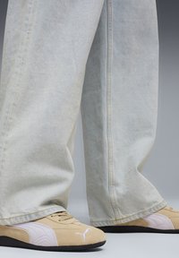 Μπεζ σουέντ sneakers με ανοιχτό ροζ λεπτομέρειες και μαύρες λαστιχένιες σόλες· φορεμένα με ανοιχτό μπλε τζιν παντελόνι με φαρδιά πόδια. Ορατές ραφές.