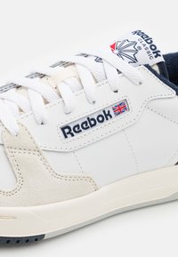 Baskets classiques blanches Reebok avec lacets blancs, détails en daim crème, et étiquette avec le logo Reebok et le drapeau britannique sur le côté.