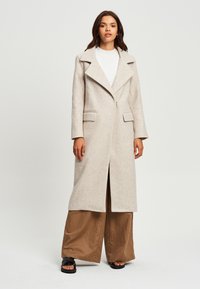 Cappotto in misto lana beige con un design doppiopetto, colletto a punte e tasche laterali; abbinato a pantaloni marroni larghi e sandali piatti.