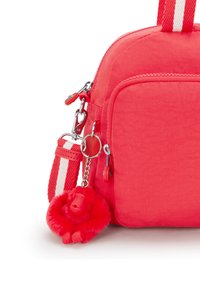 Röd tyg crossbody-väska med en dragkedjeficka, randig axelrem och en röd fluffig apa nyckelring. Texturerad yta med släta accenter.