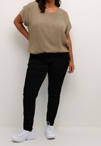 Femme se tenant debout avec une main sur la hanche, portant un haut loose à manches courtes olive, un pantalon noir et des baskets blanches sur un fond uni.