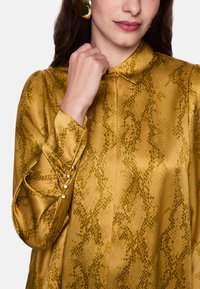 Gold snake-print blouse with a collared neckline, pearl button accents on cuffs, and smooth, glossy fabric texture.