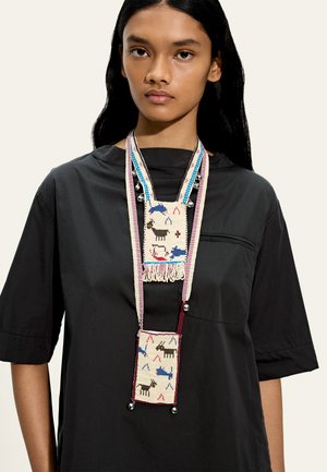 Młoda kobieta nosząca czarną koszulę z długim naszyjnikiem z koralików z motywami zwierzęcymi i geometrycznymi w kolorach niebieskim, czerwonym, czarnym i białym.