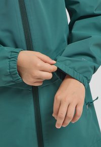 Giacca impermeabile verde acqua con chiusura a zip e polsini elasticizzati. Mani che regolano il polsino. Il tessuto appare liscio con una finitura opaca.