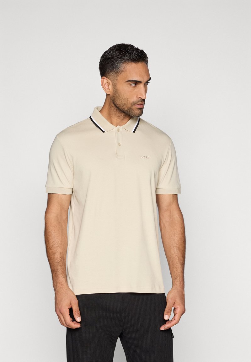 Muž v béžovém polokošili s krátkým rukávem a černobílým proužkovaným límcem, hledící doprava, stojící na jednobarevném světlém pozadí.