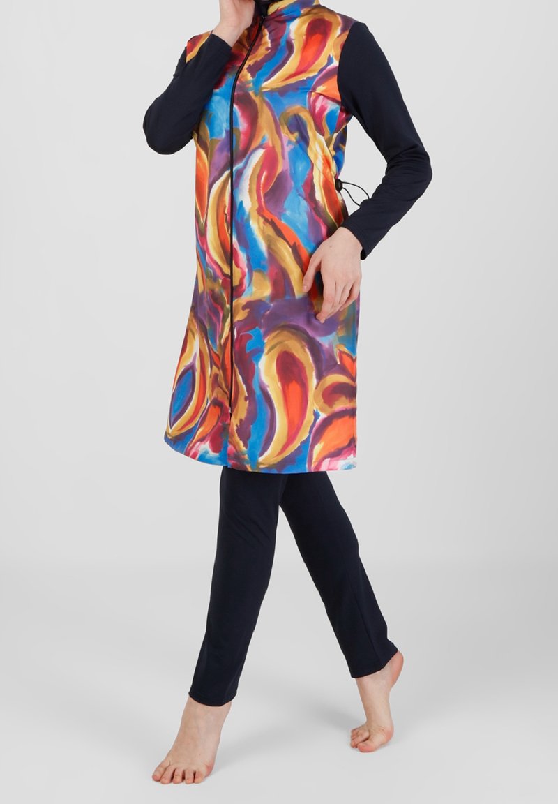 Personne portant une tunique sans manches colorée à motif abstrait par-dessus un haut noir à manches longues et des leggings noirs, debout pieds nus.