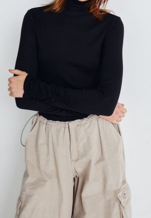 Schwarzes, geripptes Turtleneck-Oberteil mit langen Ärmeln und gerafften Manschetten, kombiniert mit hellbeigen, weit geschnittenen Cargo-Hosen. Dezente Textur sichtbar.