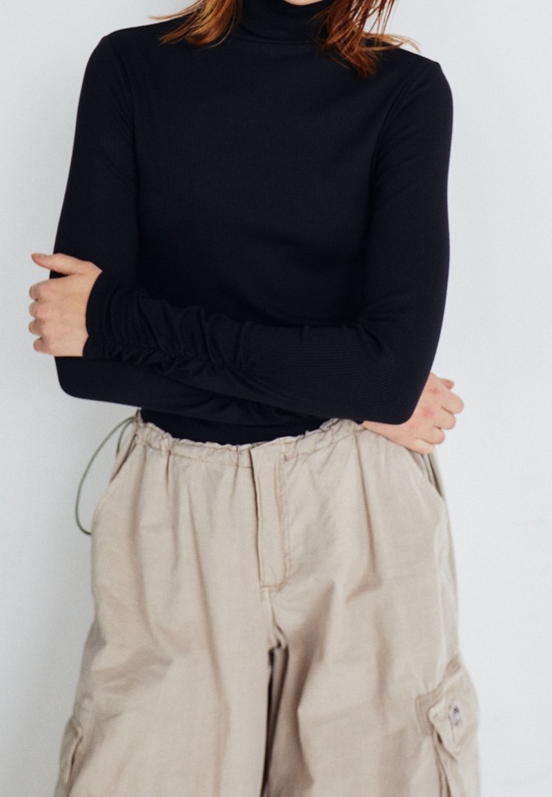Schwarzes, geripptes Turtleneck-Oberteil mit langen Ärmeln und gerafften Manschetten, kombiniert mit hellbeigen, weit geschnittenen Cargo-Hosen. Dezente Textur sichtbar.