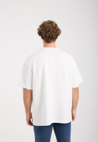 Wit oversized T-shirt met korte mouwen, gemaakt van zacht materiaal. Heeft een ronde halslijn en een ontspannen pasvorm, gecombineerd met een blauwe spijkerbroek.