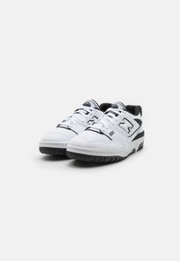 Pari di scarpe da ginnastica New Balance 550 bianche e nere a basso profilo con lacci, loghi sulle linguette e sui lati, e suole nere su uno sfondo bianco.
